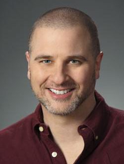 Headshot of Greg Weisman smiling casually. He is a white man with a full head of very short hair and a short beard with bits of gray on its edges. 