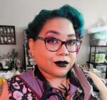 Samantha Rei headshot. She is a lighter-complexion Black woman with teal hair, dark lipstick, pierced lip and nose, and glasses. She's holding a giant pair of scissors.