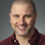 Headshot of Greg Weisman smiling casually. He is a white man with a full head of very short hair and a short beard with bits of gray on its edges.