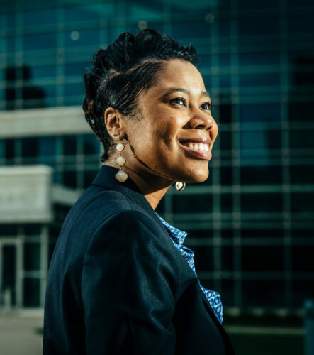 Young Black woman with styled black hair, large earrings, wearing a blazer, looks in the distance and smiles aspirationally.