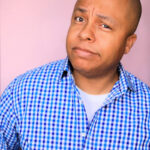 Bald Black man in a blue checkered shirt stands against a pink background, raising an eyebrow with a skeptical expression, conveying playful curiosity or doubt.