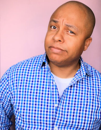 Bald Black man in a blue checkered shirt stands against a pink background, raising an eyebrow with a skeptical expression, conveying playful curiosity or doubt.