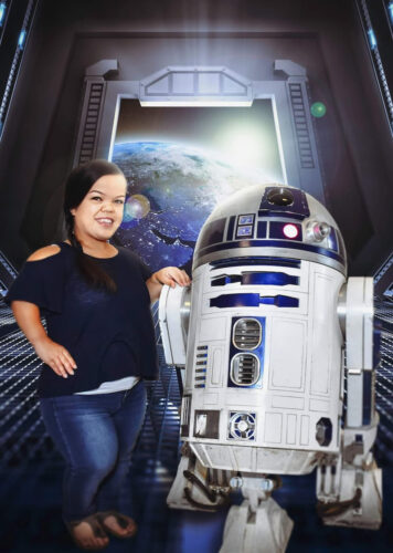 A woman with dwarfism smiles while posing with R2-D2 in a futuristic space station. Earth is visible through a large window, adding a sci-fi atmosphere.