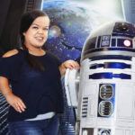 A woman with dwarfism smiles while posing with R2-D2 in a futuristic space station. Earth is visible through a large window, adding a sci-fi atmosphere.