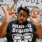 A Black man with short dreadlocks holds up glasses with both hands, making an excited face. He wears a graphic t-shirt with bold lettering that says "NUR-D Club". The background has playful, abstract doodles.
