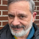 Man with gray hair and beard smiling slightly in front of a brick wall, wearing a blue hoodie and black jacket, projecting a warm and approachable demeanor.