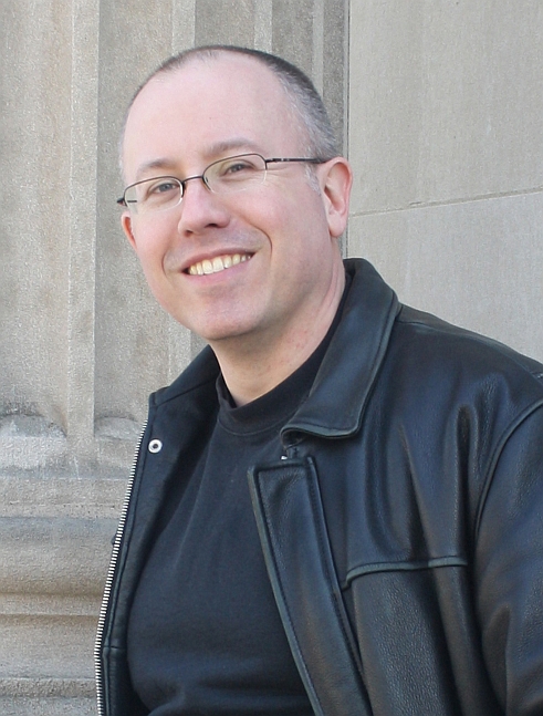 Photo of Charles Urbach, a middle-aged white man wearing glasses and a leather coat. He is smiling.
