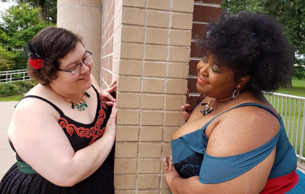 Photo of Jessica Walsh and Brianna Lawrence peeking around a wall smiling at each other. Jessica is a white-presenting plus-sized woman with a red flower in her brown hair and glasses. Briana is a plus-sized Black woman with big hair and green eye shadow. She is wearing a teal dress. Both women are wearing matching necklaces in different colors.