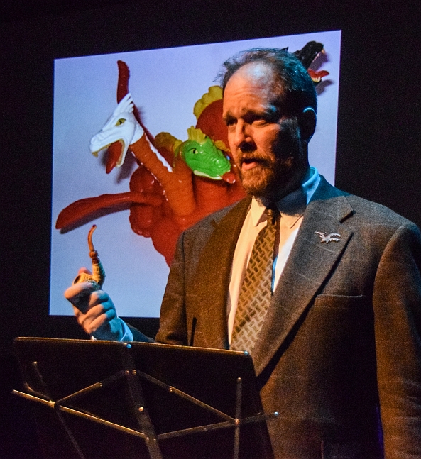 Photo of Matthew Kessen giving a monster lecture. He is a white man with brown hair and a beard. He is wearing a suit and holding a pipe like a stereotypical college professor.
