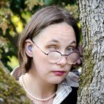 Headshot of Elsa Sjunneson peeking from behind a tree. Her hearing aid is visible in her ear and one her iris and pupil is milky white on one eye. Her other eye is blue. She is wearing purple glasses and pearls and has short, brown hair.