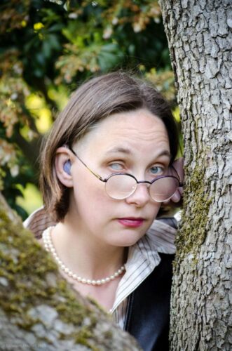 Headshot of Elsa Sjunneson peeking from behind a tree. Her hearing aid is visible in her ear and one her iris and pupil is milky white on one eye. Her other eye is blue. She is wearing purple glasses and pearls and has short, brown hair.