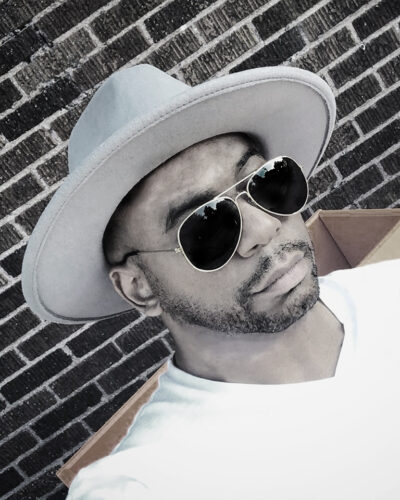 Headshot of T. Aaron Cisco. He is a Black man in dark sunglasses with light facial hair, smiling slightly at the camera, wearing a gray hat.
