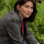 Headshot of Laura Anne Gilman. She is wearing jeans and a blazer and smiling coyly at the camera. Her glasses are hanging from her collar. She has dark brown, shoulder-length hair.