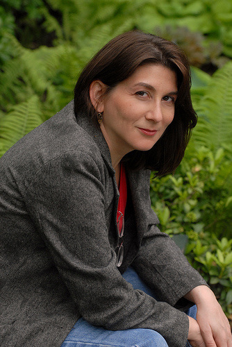 Headshot of Laura Anne Gilman. She is wearing jeans and a blazer and smiling coyly at the camera. Her glasses are hanging from her collar. She has dark brown, shoulder-length hair.