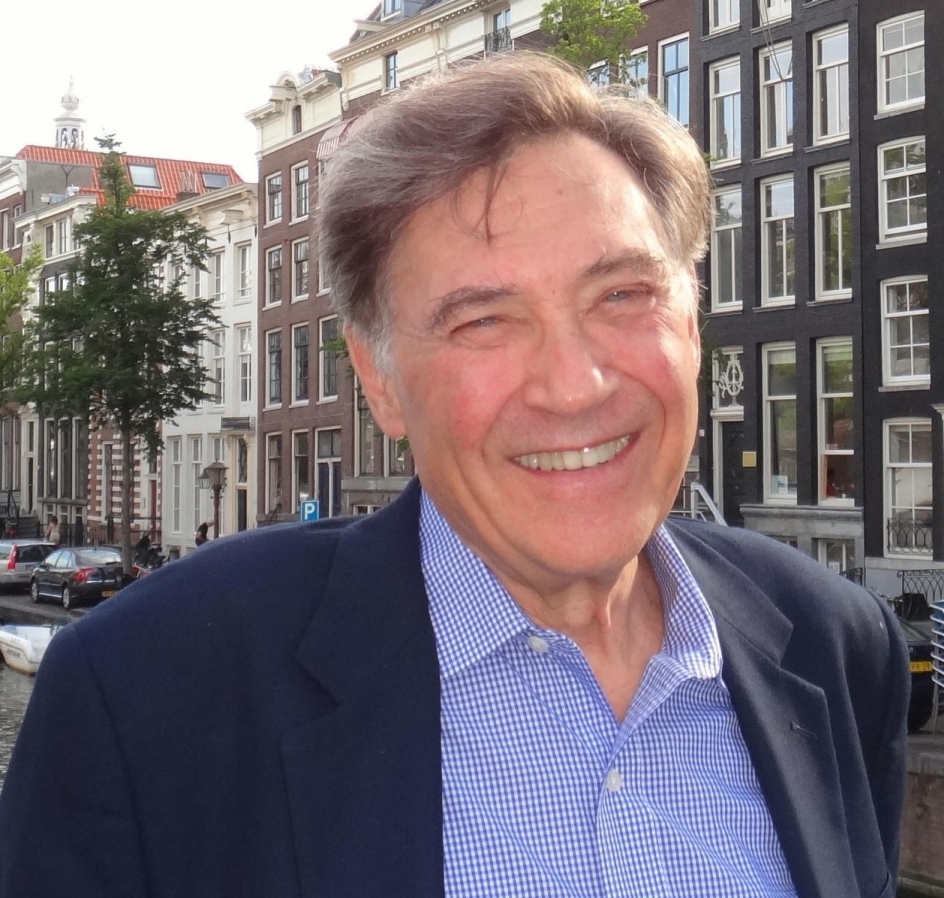 Photo of Jack Zipes. He is an older white man with brown hair, slightly bushy eyebrows, blue eyes with a twinkle in them. He is smiling, wearing a blue wool coat, with the back drop of a European city street behind him.