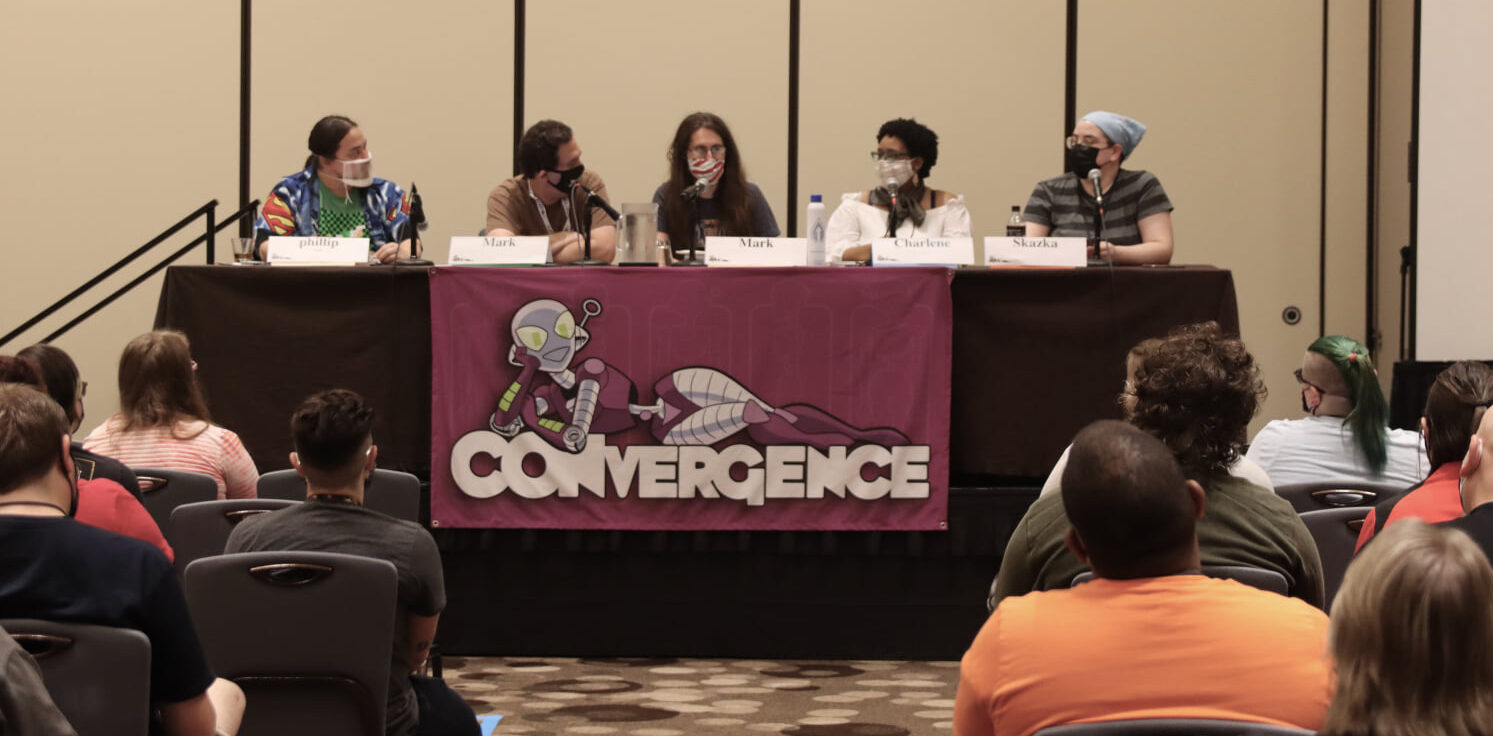 Panelists wearing face masks at a table with a CONvergence banner on it at CONvergence 2022