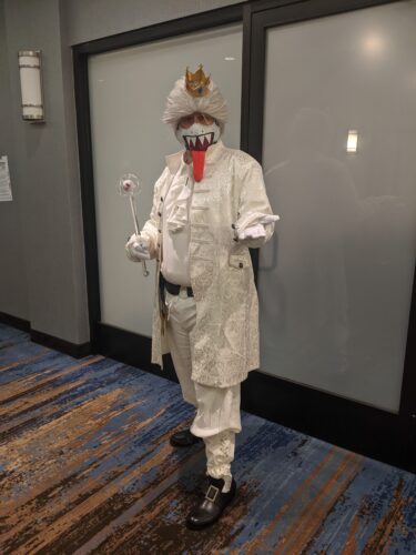 CONvergence 2021 cosplayer in a white regency suit, white wig with little crown, and face mask with a big sharp-toothed smile and tongue on it