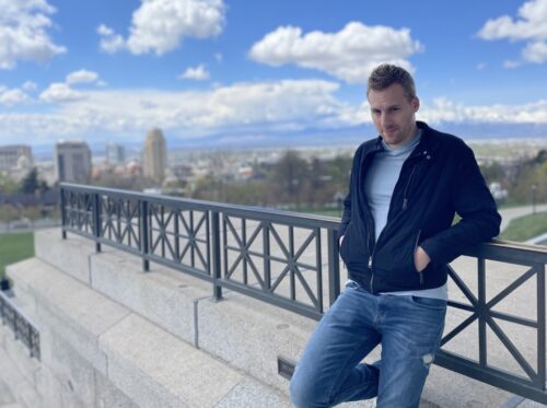 Photo of Theology, a white male-presenting person in ripped jeans, leaning on a railing in front of a city
