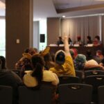 Audience at a panel, one person is holding up an ASL I love you sign