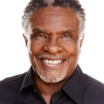 Headshot of Keith David. He is an older Black man with short white goatee-style facial hair and streaks of white hair at his temples and on the top of his short curly hair. He is smiling warmly, with twinkling eyes.