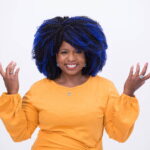 Miss Shannan Paul smiles, holding hands in front of her making an inviting gesture. She is a Black woman with large curly black hair with blue streaks throughout it. She is wearing very bright colors and strikingly-contrasting jewelry.