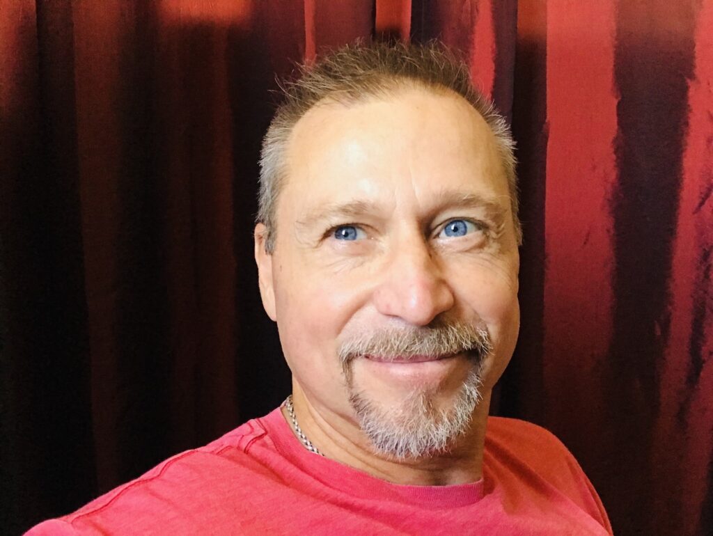 Selfie of Thom Adcox smiling. He is a white man with short hair and a goatee, both peppered with gray. He has sparkling blue eyes.