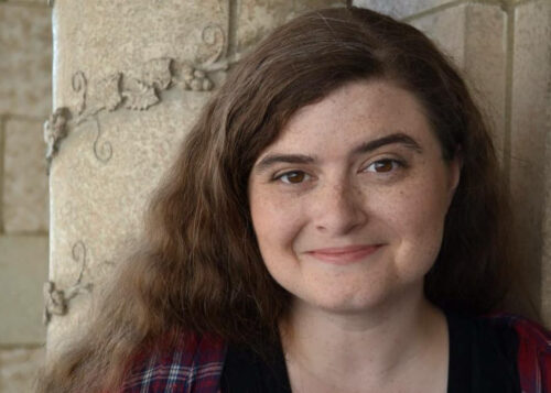 A woman with long brown hair and a slight smile stands against a textured stone wall with vine engravings, conveying warmth and friendliness.