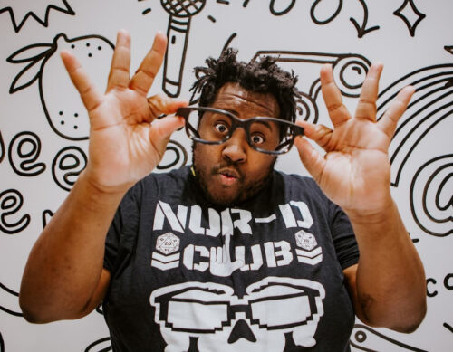 A Black man with short dreadlocks holds up glasses with both hands, making an excited face. He wears a graphic t-shirt with bold lettering that says "NUR-D Club". The background has playful, abstract doodles.
