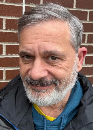Man with gray hair and beard smiling slightly in front of a brick wall, wearing a blue hoodie and black jacket, projecting a warm and approachable demeanor.